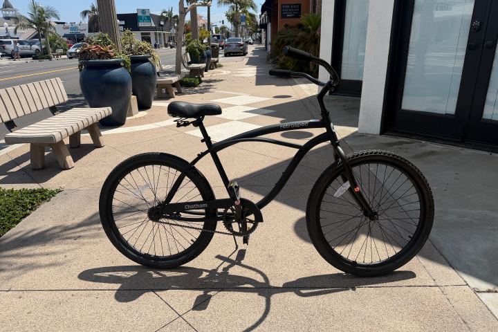 Black bicycle parked on a sunny sidewalk near benches and potted plants.