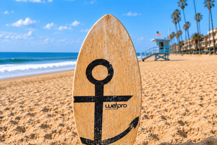 Surfboard with anchor design on sandy beach, palm trees and lifeguard tower in background.