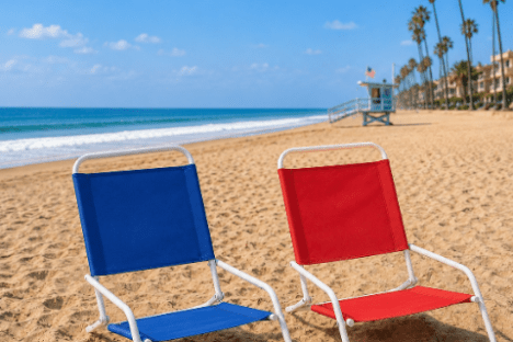 Two beach chairs on sand, palm trees and ocean in background, blue sky.