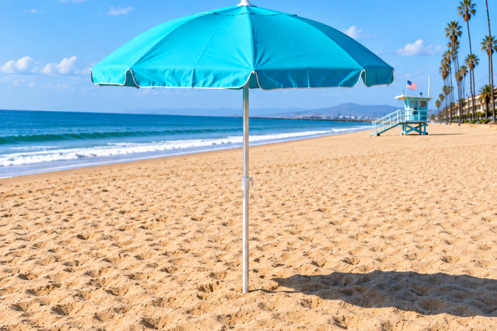 Blue beach umbrella on sandy beach with ocean and palm trees in background.